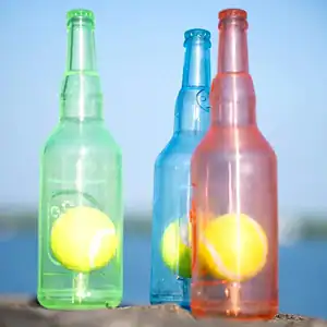 Three clear bottles—green, blue, pink—each hold a yellow tennis ball against a blurred outdoor backdrop.