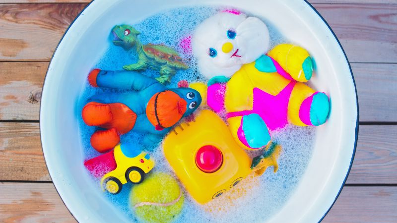Cleaning toys in a soapy water tub on a wooden floor, including plush animals, a toy car, and a rubber duck.