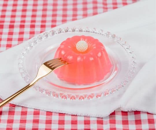 Postre de gelatina rosa con una perla en la parte superior, servido en un plato de vidrio con un tenedor dorado sobre una servilleta sobre un mantel a cuadros rojos.