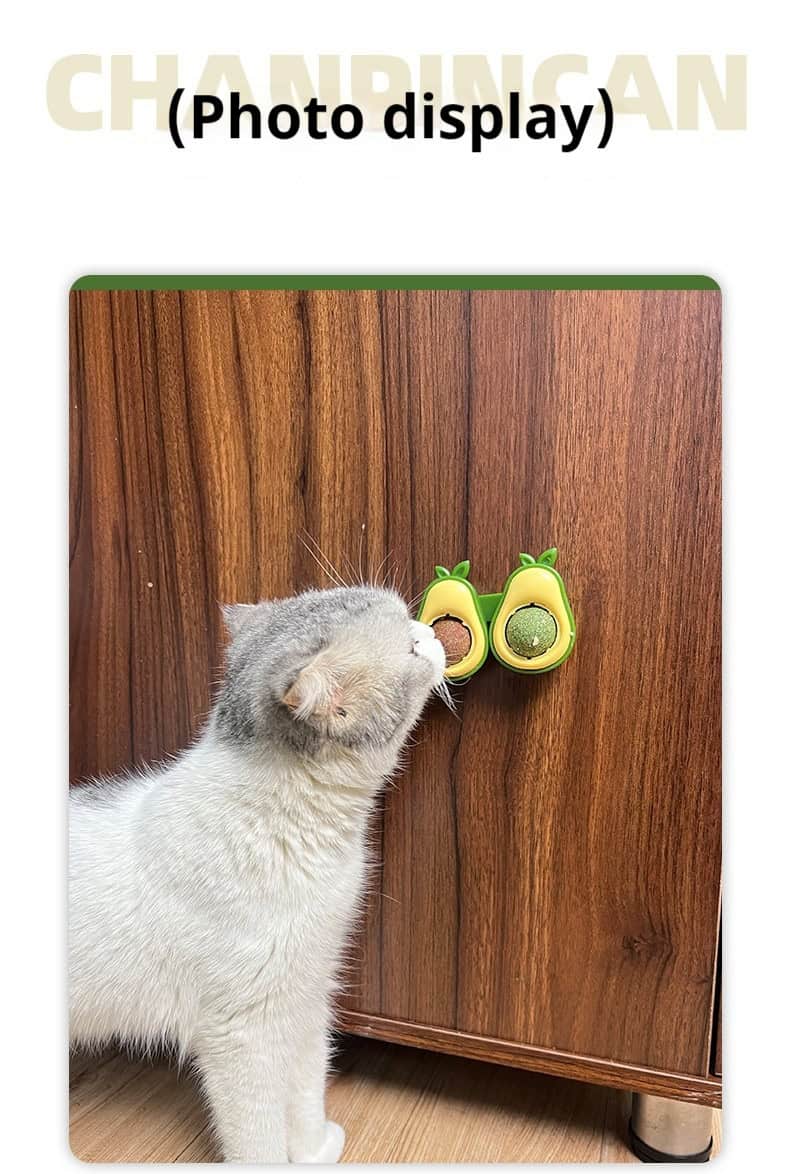A white and gray cat sniffs an avocado-shaped catnip toy attached to a wooden cabinet.