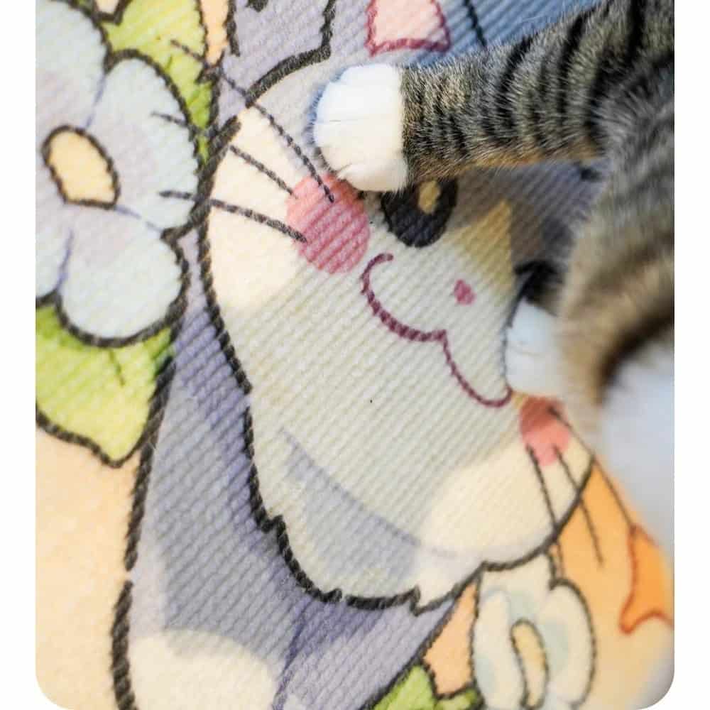 A cat's front paws step on a colorful cartoon cat-face scratching pad with floral designs.