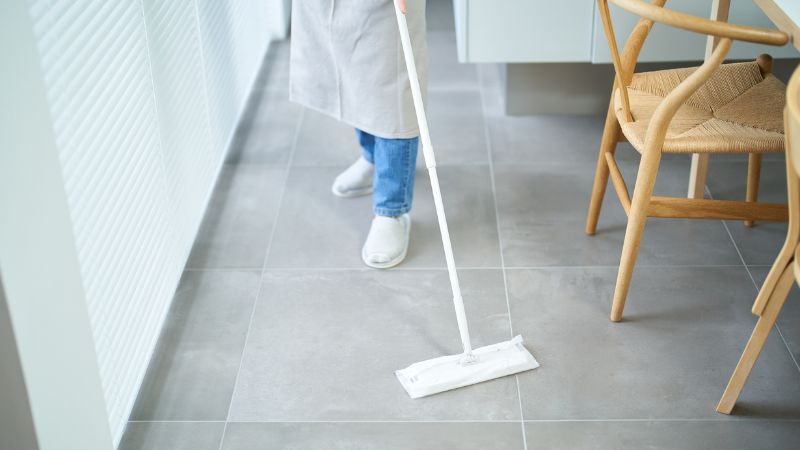Freshly mopped gray tiled floor in a modern kitchen or dining area with a person cleaning with a mop, emphasizing cleanliness and hygiene.