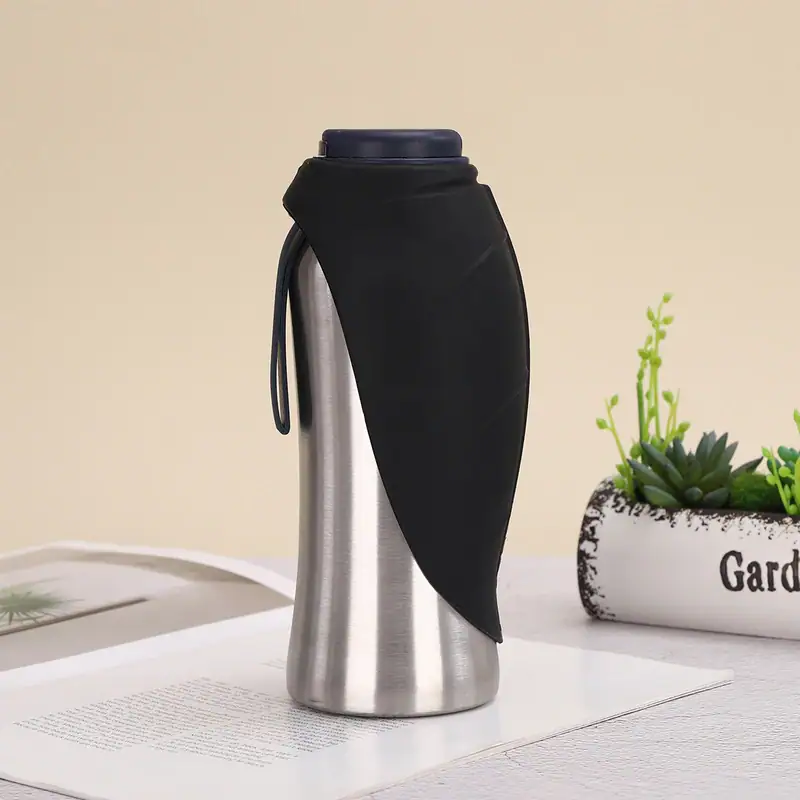 Stainless steel travel mug with black silicone cover and lid, placed on a white surface with a plant in the background.