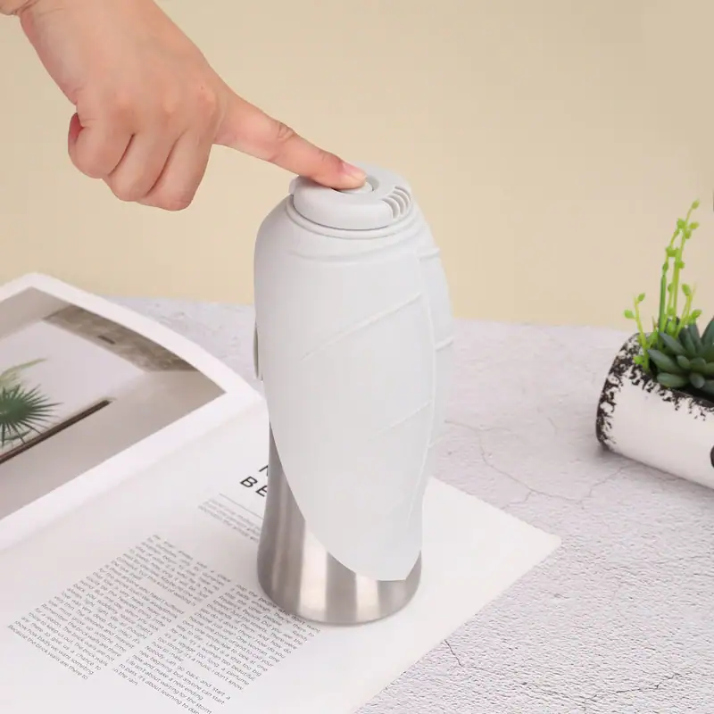 Close-up of a white portable travel mug with a button lid, placed on a table with a magazine and a small potted plant nearby.